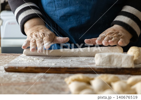 An elderly person shapes soft dough on a floured wooden board to create handmade dumplings, reflecting a traditional culinary process, authentic cooking skills, and home food culture An elderly person shapes soft dough on a floured wooden board to create handmade dumplings, reflecting a traditional culinary process, authentic cooking skills, and home food culture 140676519