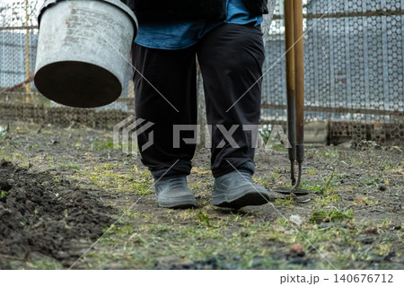 A gardener walking with a plastic bucket and a metal hoe in a backyard, embodying agricultural effort, outdoor maintenance, and sustainable lifestyle practices in a rural setting A gardener walking with a plastic bucket and a metal hoe in a backyard, embodying agricultural effort, outdoor maintenance, and sustainable lifestyle practices in a rural setting 140676712