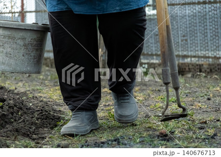 A gardener walking across a dirt backyard plot while carrying a plastic bucket and a hand hoe, demonstrating active garden maintenance during the spring season for a healthy lawn A gardener walking across a dirt backyard plot while carrying a plastic bucket and a hand hoe, demonstrating active garden maintenance during the spring season for a healthy lawn 140676713