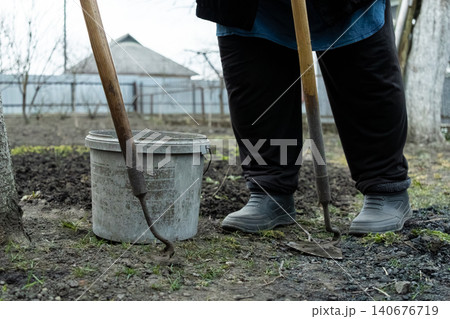 A gardener uses a pair of metal hand hoes near a plastic bucket on fresh soil to cultivate a rural vegetable plot, reflecting humble agricultural work and seasonal land care A gardener uses a pair of metal hand hoes near a plastic bucket on fresh soil to cultivate a rural vegetable plot, reflecting humble agricultural work and seasonal land care 140676719