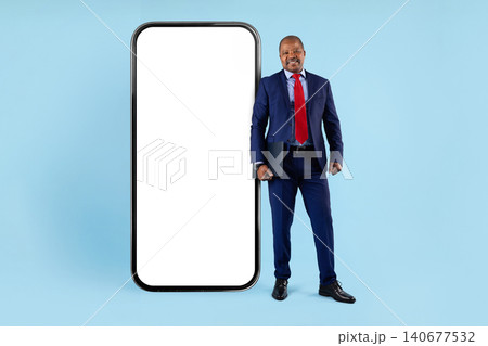 Mature black businessman in suit holding folder, standing beside giant blank smartphone Mature black businessman in suit holding folder, standing beside giant blank smartphone 140677532