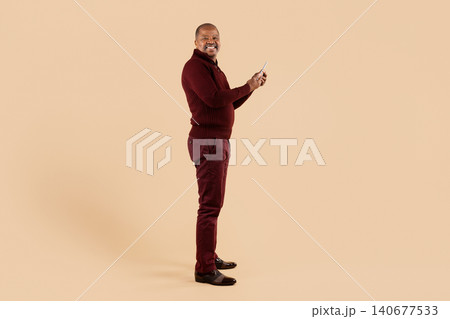 Mature black man in burgundy sweater holding smartphone while standing in profile Mature black man in burgundy sweater holding smartphone while standing in profile 140677533