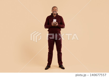 Mature African American man with smartphone in hands standing on beige background Mature African American man with smartphone in hands standing on beige background 140677557