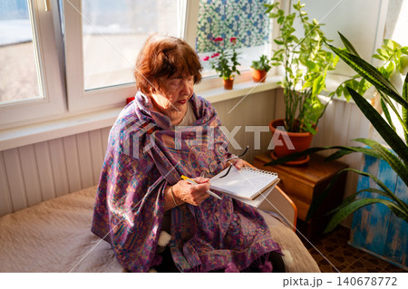 Caucasian elderly woman recording ideas in a notebook while enjoying a peaceful morning by the window. Education and creativity in retirement. 140678772