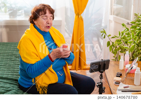Caucasian grandmother reviewing decorative cosmetics while engaging with followers during an online broadcast. Social media influencer concept. Modern retirement and creativity. 140678780