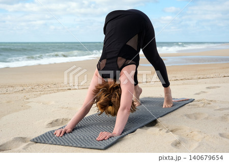 Woman practicing yoga on a sandy beach in downward facing dog pose, promoting wellness, fitness, mindfulness, healthy lifestyle, relaxation, and balance 140679654