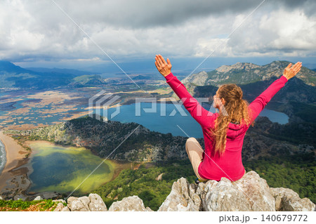 Woman sits on high cliff with raised hands overlooking Dalyan delta and Iztuzu Turtle Beach in Turkey. Inspiring travel moment symbolizing freedom, adventure and summer family tourism. 140679772