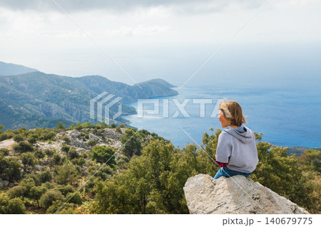Young boy sits on a rocky viewpoint overlooking the sea in Turkey. Calm travel moment with wide coastal landscape, inspiring exploration and outdoor family adventure. Young boy sits on a rocky viewpoint overlooking the sea in Turkey. Calm travel moment with wide coastal landscape, inspiring exploration and outdoor family adventure. 140679775
