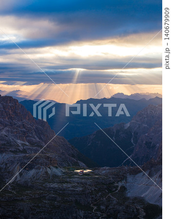 Powerful sunbeams break through dramatic storm clouds above the Tre Cime di Lavaredo region in the Dolomites, Italy. Aerial view of rugged alpine terrain illuminated by golden evening light. Powerful sunbeams break through dramatic storm clouds above the Tre Cime di Lavaredo region in the Dolomites, Italy. Aerial view of rugged alpine terrain illuminated by golden evening light. 140679909