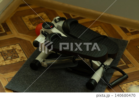White stepper machine with black pedals and hydraulic piston on a grey mat for home fitness training. White stepper machine with black pedals and hydraulic piston on a grey mat for home fitness training. 140680470