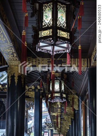 An ornate, hexagonal lantern hangs from a dark wooden ceiling. Delicate, glowing glass panels are framed by intricate woodwork. Long, vibrant red tassels dangle from each corner. An ornate, hexagonal lantern hangs from a dark wooden ceiling. Delicate, glowing glass panels are framed by intricate woodwork. Long, vibrant red tassels dangle from each corner. 140680533