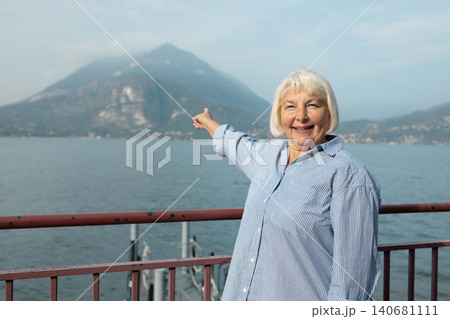 Joyful smiling mature elderly caucasian female traveler with backpack standing on promenade and pointing away towards scenic lake and mountains. Joyful smiling mature elderly caucasian female traveler with backpack standing on promenade and pointing away towards scenic lake and mountains. 140681111