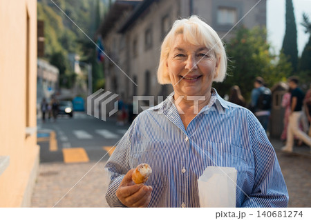 Happy senior woman tourist holding paper bag with pastry outdoors Happy senior woman tourist holding paper bag with pastry outdoors 140681274