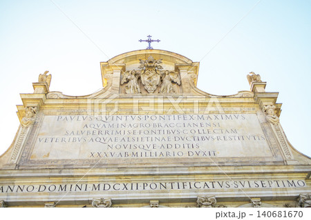 Close-up of ancient marble attic with historical Latin inscriptions, papal coat of arms and cross against blue sky. Close-up of ancient marble attic with historical Latin inscriptions, papal coat of arms and cross against blue sky. 140681670