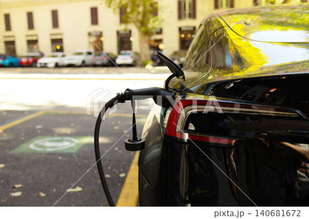 Close-up of electric car charging plug inserted into power station connector with vehicles in background. Close-up of electric car charging plug inserted into power station connector with vehicles in background. 140681672