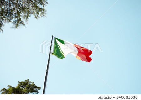 Italian flag tricolor waving in the wind against a bright blue sky with green trees. Italian flag tricolor waving in the wind against a bright blue sky with green trees. 140681688