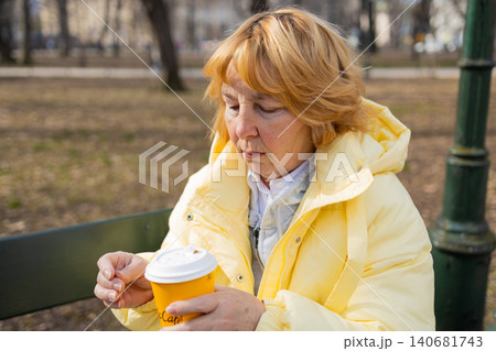 Mature woman in a yellow jacket drinking coffee on a park bench. Mature woman in a yellow jacket drinking coffee on a park bench. 140681743