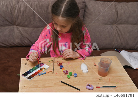 Young girl decorating white plaster figurine at wooden table using colorful paints. 140681791