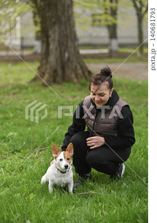 Woman kneeling beside Jack Russell dog on green grass in park. Woman kneeling beside Jack Russell dog on green grass in park. 140681793