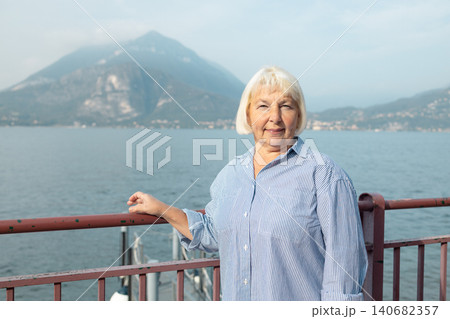 Joyful smiling mature elderly caucasian female traveler with backpack standing on promenade and pointing away towards scenic lake and mountains. Joyful smiling mature elderly caucasian female traveler with backpack standing on promenade and pointing away towards scenic lake and mountains. 140682357