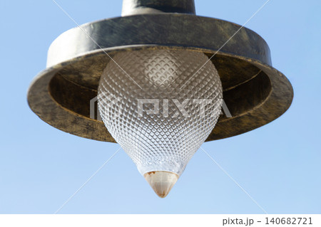 Streetlight lamp glass bulb outdoor fixture vintage metal, closeup lighting detail with textured shade and diffuser, hanging overhead against blue sky background, calm minimal exterior Streetlight lamp glass bulb outdoor fixture vintage metal, closeup lighting detail with textured shade and diffuser, hanging overhead against blue sky background, calm minimal exterior 140682721