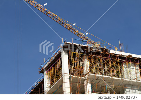 Construction site crane scaffolding concrete building highrise skyline under clear blue sky, ambitious urban development with exposed framework and safety netting Construction site crane scaffolding concrete building highrise skyline under clear blue sky, ambitious urban development with exposed framework and safety netting 140682737