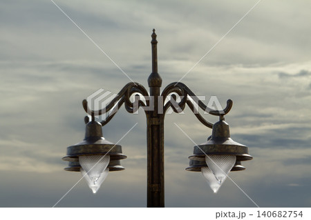 Streetlight lamp lantern metal vintage ornate pole sky, twin outdoor light fixture with wrought iron scrollwork against cloudy evening horizon, urban lighting detail 140682754