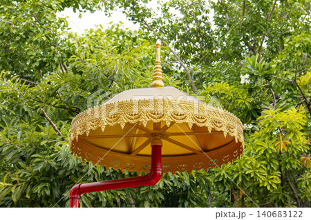 Golden parasol with ornate trim above red post in lush green garden, decorative canopy in outdoor nature setting Golden parasol with ornate trim above red post in lush green garden, decorative canopy in outdoor nature setting 140683122