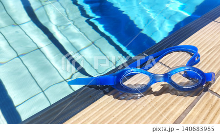 Blue swimming goggles resting on the edge of a clear outdoor pool, reflecting summer leisure, fitness, and vacation vibes under bright sunlight. Blue swimming goggles resting on the edge of a clear outdoor pool, reflecting summer leisure, fitness, and vacation vibes under bright sunlight. 140683985