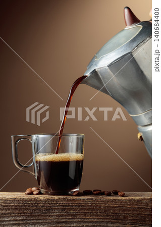 Black coffee is poured from a classic Italian aluminium moka pot into a glass coffee cup. Black coffee is poured from a classic Italian aluminium moka pot into a glass coffee cup. 140684400