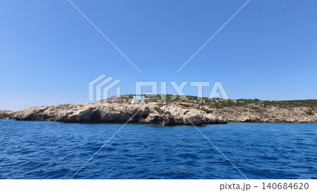 The island's rocky coastline, surrounded by crystal-clear sea and a clear sky, features a small white house with a blue roof. Mediterranean landscape with coastal cliffs and calm waters. The island's rocky coastline, surrounded by crystal-clear sea and a clear sky, features a small white house with a blue roof. Mediterranean landscape with coastal cliffs and calm waters. 140684620