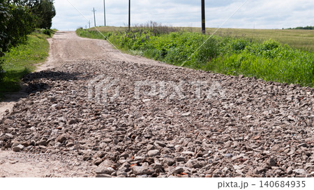 A rough gravel road stretches through a quiet rural landscape with green grass, utility poles, and open fields under a bright summer sky A rough gravel road stretches through a quiet rural landscape with green grass, utility poles, and open fields under a bright summer sky 140684935