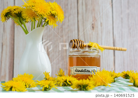 Transparent glass of jar dandelion jelly wooden honey dipper coated with jelly dripping with dandelion flowers in vase. Soft natural lighting highlights the amber color glossy texture and artisanal Transparent glass of jar dandelion jelly wooden honey dipper coated with jelly dripping with dandelion flowers in vase. Soft natural lighting highlights the amber color glossy texture and artisanal 140691525