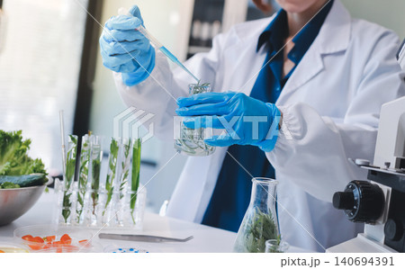 Scientists in lab coats work with plants in a modern lab, focusing on research and experimentation under bright, sterile conditions. 140694391
