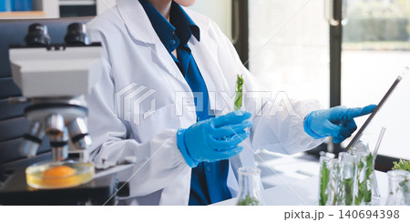 Scientists in lab coats work with plants in a modern lab, focusing on research and experimentation under bright, sterile conditions. 140694398