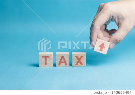 A close-up of a person's hand placing a wooden cube with an upward arrow next to the word tax on a blue background. High Quality Photo 140695494