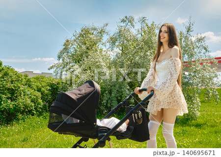 Cute mother walks in park with stroller on sunny day surrounded by green trees and grass Cute mother walks in park with stroller on sunny day surrounded by green trees and grass 140695671