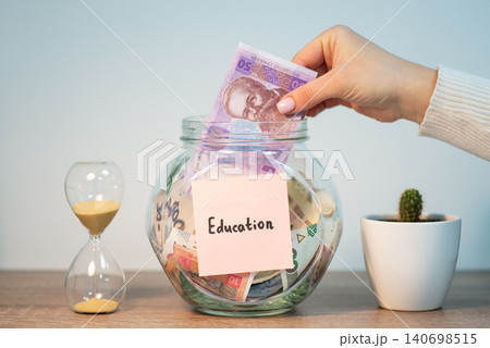 Female hand putting 50 hryvnia banknote to glass jar with inscription education. Saving up money for education in Ukraine Female hand putting 50 hryvnia banknote to glass jar with inscription education. Saving up money for education in Ukraine 140698515