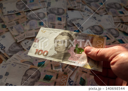 One hundred hryvnia banknote in mans hand, 500 hryvnia banknotes on the background. Ukrainian hryvnias in cash, big sum of money on the table One hundred hryvnia banknote in mans hand, 500 hryvnia banknotes on the background. Ukrainian hryvnias in cash, big sum of money on the table 140698544