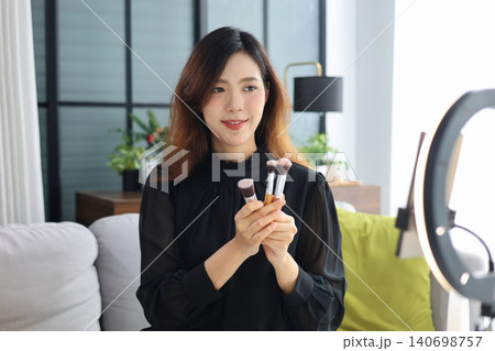 Beautiful Asian beauty influencer holding makeup brushes during online live stream content creation at home. 140698757