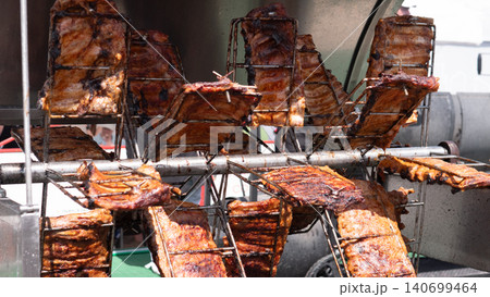 Several slabs of meat are cooking on metal racks over an open grill setup, with dark charred surfaces, warm browning, and industrial cooking equipment visible in the background Several slabs of meat are cooking on metal racks over an open grill setup, with dark charred surfaces, warm browning, and industrial cooking equipment visible in the background 140699464