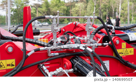 Close-up view of red industrial machinery with multiple hydraulic hoses, metal connectors, and safety labels, set outdoors with a blurred background and natural daylight 140699507
