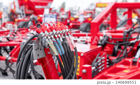Close-up view of red farm equipment showing bundled hydraulic hoses, metal connectors, and industrial components with a blurred machinery background and bright outdoor lighting 140699551