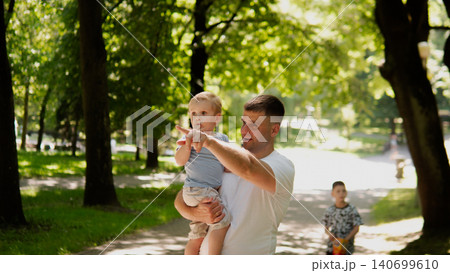 Young father holding toddler boy at shoulder height while pointing ahead along sunlit tree-lined park path; dappled afternoon light filters through leafy 140699610