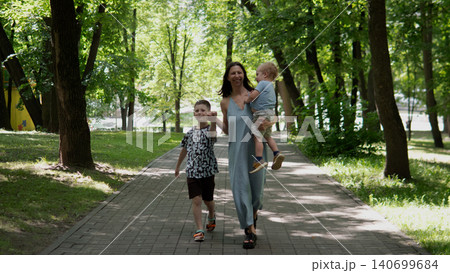 Woman wearing a light summer dress walks down a tree-lined paved path in a green park, holding a toddler in one arm while an older child walks beside her Woman wearing a light summer dress walks down a tree-lined paved path in a green park, holding a toddler in one arm while an older child walks beside her 140699684