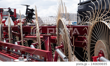 Close view of a red farm implement with multiple large metal wheels and curved tines parked outdoors under a bright sky, showing mechanical parts, a steel frame, and rugged rural Close view of a red farm implement with multiple large metal wheels and curved tines parked outdoors under a bright sky, showing mechanical parts, a steel frame, and rugged rural 140699695