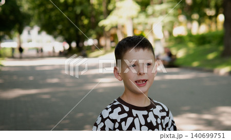 Boy around seven standing on a tree-lined park path, smiling with slightly open mouth, wearing patterned dark and white shirt 140699751