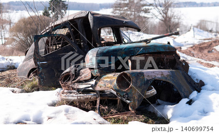 Heavy rusted vintage car wreck sits abandoned in melting snow within rural landscape, torn roof and crushed front end revealing corroded metal panels, broken 140699754
