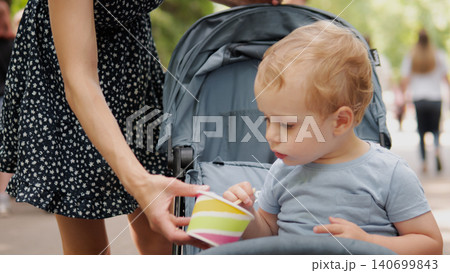 Curious toddler seated in a stroller reaches for a striped paper cup offered by a standing parent wearing a black floral dress; soft natural daylight filters Curious toddler seated in a stroller reaches for a striped paper cup offered by a standing parent wearing a black floral dress; soft natural daylight filters 140699843