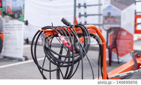 Coiled black cables and connectors hang on an orange metal stand in an indoor exhibition space, with blurred informational banners, product displays, and bright ambient lighting 140699856